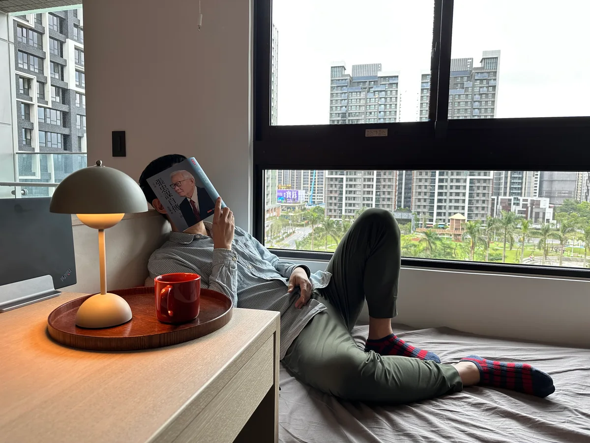 KC 在家中的窗邊閱讀，窗外是熟悉的城市街景。